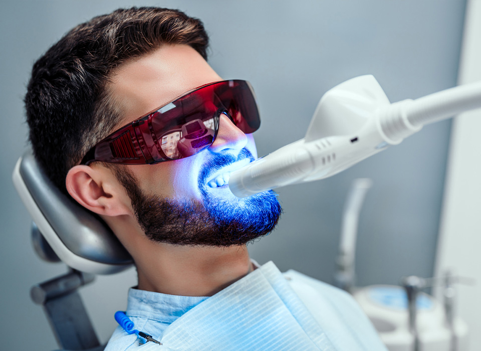 A dentist is cleaning a man's teeth in a dental office, with tools and equipment visible in the background.