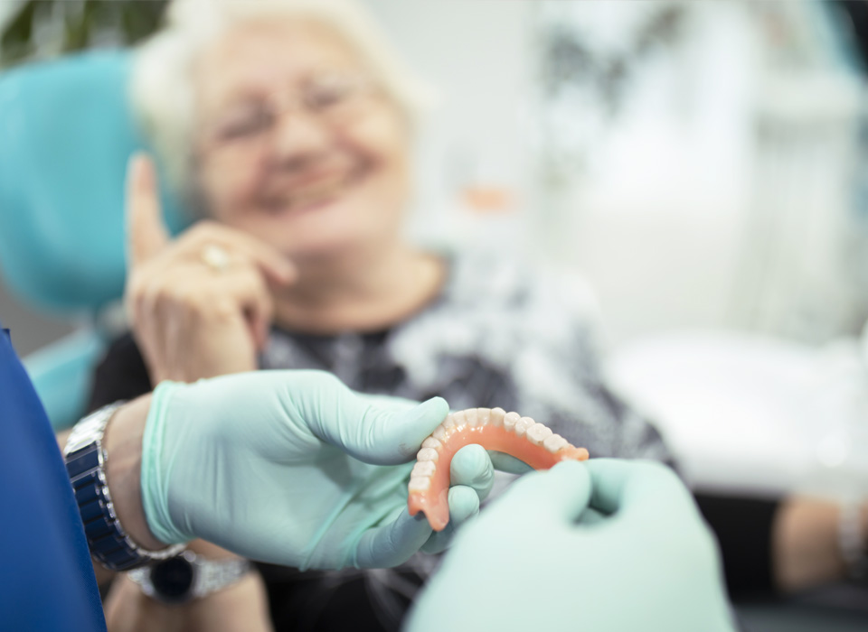A blurry image of an old lady patient with a smile on a dental chair while dentures being hold by dentist hands