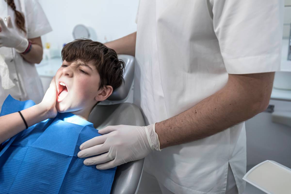 Worried looking boy in the dentist chair in front of the doctor is complaining because of her gum pain and holding her hand on the cheek.
